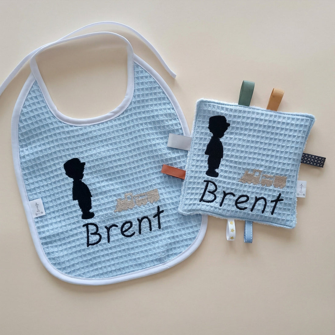 Handgemaakte kraamset voor jongens met gepersonaliseerd slabbetje en knisperdoekje van wafelstof in babyblauw, met borduur van jongetje en trein.
