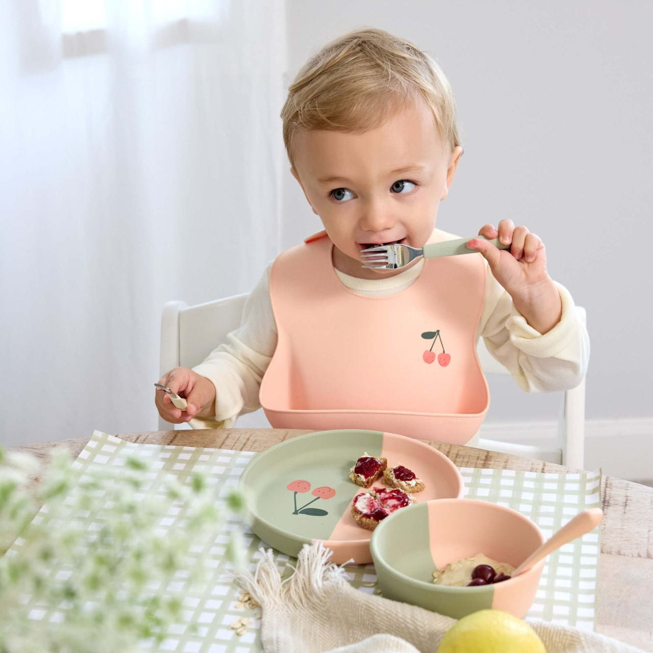 kind draagt de Lässig Happy Fruits Kersen siliconen slab aan tafel – diepe opvangzak vangt gemorst eten op